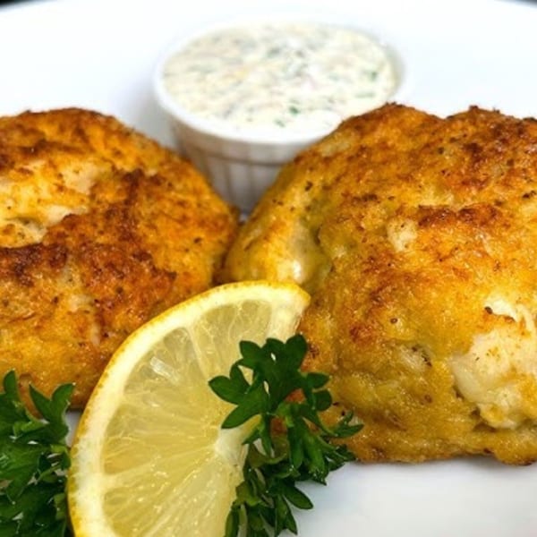 Two golden-brown crab cakes with a lemon wedge, parsley, and a side of tartar sauce.