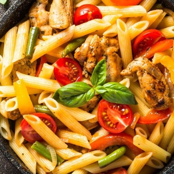 Penne pasta with grilled chicken, cherry tomatoes, green beans, and basil in a black pan.