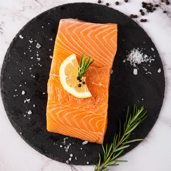 Raw salmon fillet with lemon, rosemary, salt, and peppercorns on a black slate board.