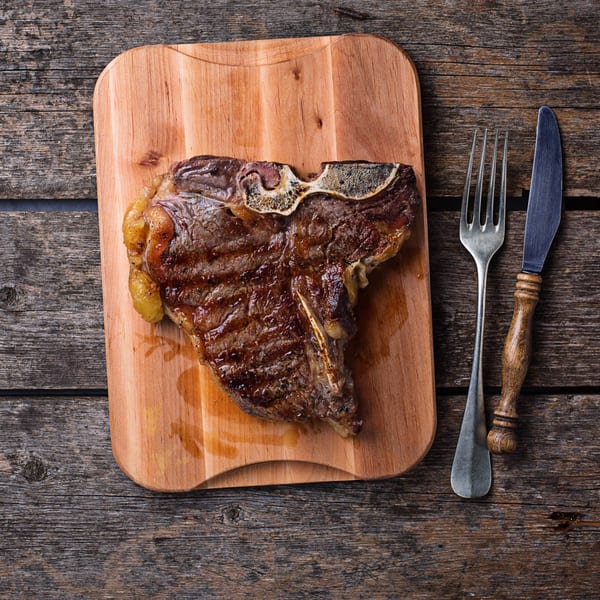 Grilled T-bone steak on a wooden cutting board with a fork and knife.